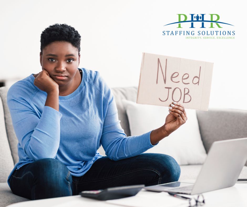 woman of color holding sign saying "need job" in front of laptop computer. Looking frustrated and disappointed.
